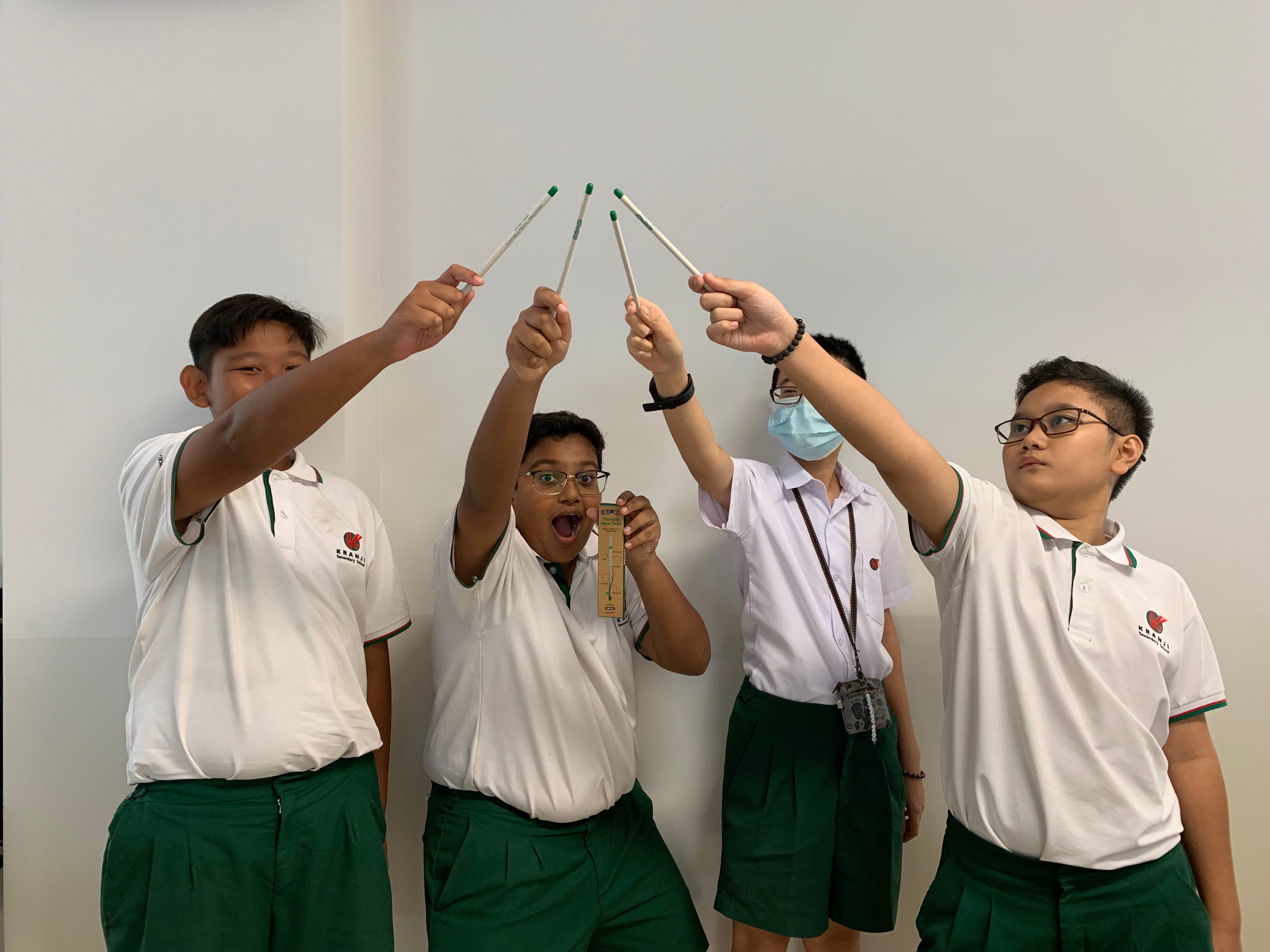 Four students in white and green uniforms raise drumsticks overhead, one wearing a mask, another holding a small box.